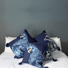 Load image into Gallery viewer, Black cockatoo and sage grevillea printed on indigo square linen designer cushion with tassels on white sheets on bed in bedroom with  charcole walls

