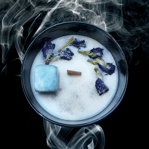 A picture of a large white candle sprinkled with butterfly pea  petals and a  Aquamarine  crystal imbedded in it